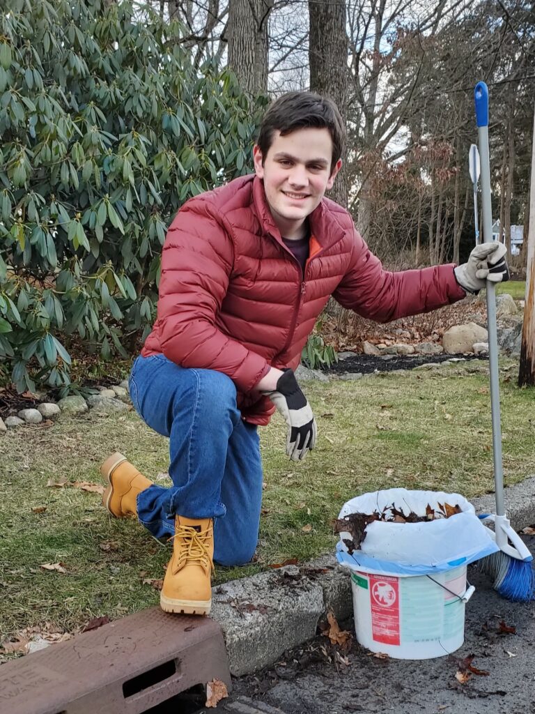 Liam Curran kneels by a storm drain that he's cleaning with a broom.